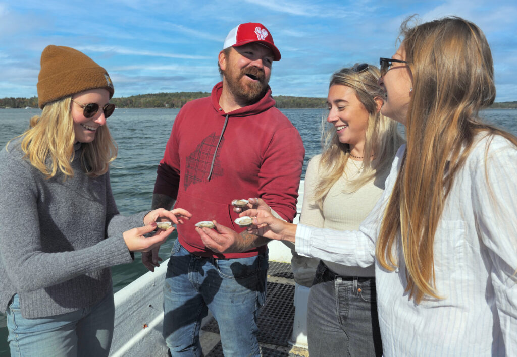 A group of friends toasting oysters on a skiff in Maine on the water. This new collection of resources from Maine Sea Grant features an Oyster Guide.